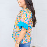 Woman with dark hair standing in side profile position wearing the Bell Top against a white background. The focal point of this top is to see the length of the top, side graphics, and ruffles on sleeve. The model paired this top with dark-wash denim shorts.
