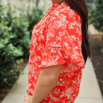 Woman with dark hair standing in a side profile position wearing the Bell Top in a lifestyle scene. The focal point of this image is the ruffled sleeves and white floral on red fabric. The model paired this top with light-wash denim jeans. 