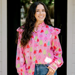 Woman with dark hair wearing a multi-color Christmas tree graphic long-sleeve with ruffles on sleeves. Model paired this top with dark-wash denim jeans. 