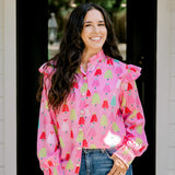 Woman with dark hair wearing a multi-color Christmas tree graphic long-sleeve with ruffles on sleeves. Model paired this top with dark-wash denim jeans. 
