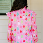 Woman with dark hair wearing a multi-color Christmas tree graphic long-sleeve with ruffles on sleeves. Model paired this top with dark-wash denim jeans. 