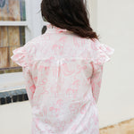 Woman with dark hair standing in a back profile position wearing the Bridget Top against a white background. the focal point of this image is the pink western toile graphics with ruffles on sleeve. The model paired this top with light-wash denim jeans. 