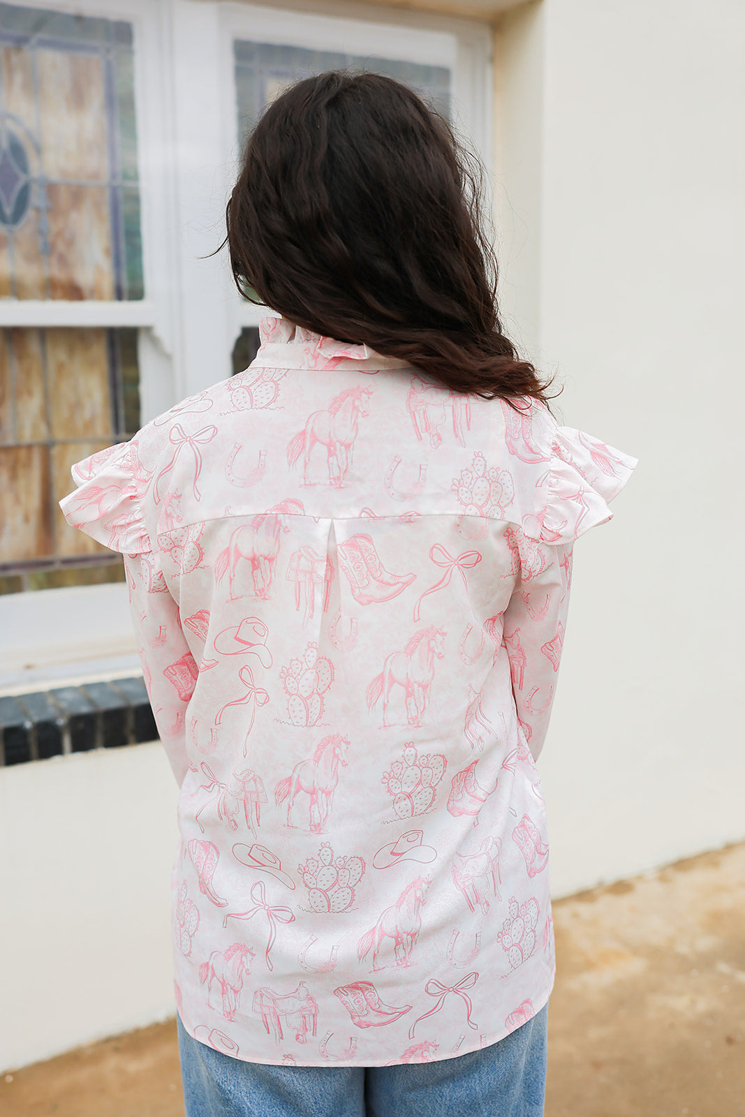 Woman with dark hair standing in a back profile position wearing the Bridget Top against a white background. the focal point of this image is the pink western toile graphics with ruffles on sleeve. The model paired this top with light-wash denim jeans. 