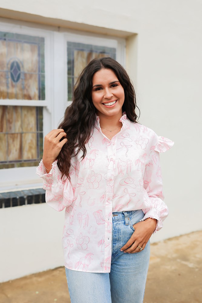 Woman with dark hair standing in a side profile position wearing the Bridget Top against a white background. The focal point of this image is the front buttons as well as the western toile graphics and ruffles on sleeve. The model paired this top with light-wash denim jeans. 