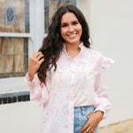 Woman with dark hair standing in a side profile position wearing the Bridget Top against a white background. The focal point of this image is the front buttons as well as the western toile graphics and ruffles on sleeve. The model paired this top with light-wash denim jeans. 