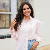 Woman with dark hair standing in a side profile position wearing the Bridget Top against a white background. The focal point of this image is the front buttons as well as the western toile graphics and ruffles on sleeve. The model paired this top with light-wash denim jeans. 