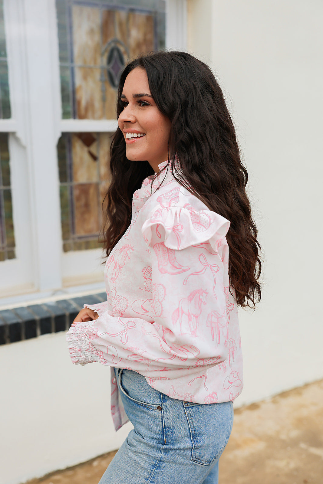 Woman with dark hair standing in a side profile position wearing the Bridget Top against a white background. The focal point of this image is the western toile graphics and ruffles on sleeve. The model paired this top with light-wash denim jeans. 