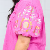 Woman with dark hair standing in a side profile position with a deep focal length. The focus of this shot is on the shoulder/sleeve region, emphasizing the embroidery on the top's sleeve. 