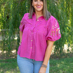 Woman with short, dark hair standing in a lifestyle scene wearing the Brinley Top. The background of model has heavy greenery, and the model is wearing light-wash denim jeans.