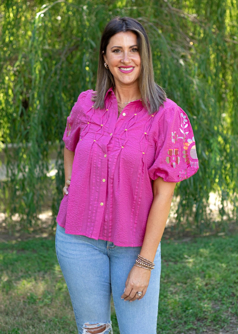 Woman with short, dark hair standing in a lifestyle scene wearing the Brinley Top. The background of model has heavy greenery, and the model is wearing light-wash denim jeans.