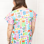 Woman with dark hair standing in a back profile position wearing the Butterfly Top against a white background. The focal point of this image is the colorful floral graphics as well as pink embroidery on back of sleeves. The model paired this top with dark-wash denim shorts. 