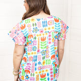 Woman with dark hair standing in a back profile position wearing the Butterfly Top against a white background. The focal point of this image is the colorful floral graphics as well as pink embroidery on back of sleeves. The model paired this top with dark-wash denim shorts. 