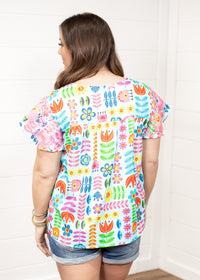 Woman with dark hair standing in a back profile position wearing the Butterfly Top against a white background. The focal point of this image is the colorful floral graphics as well as pink embroidery on back of sleeves. The model paired this top with dark-wash denim shorts. 