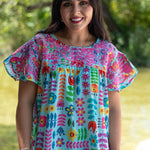 Woman with dark hair standing in a front profile position wearing the Butterfly Top in a lifestyle scene. The focal point of this image is the front detailed embroidery and floral graphics. The model paired this top with light-wash denim pants. 