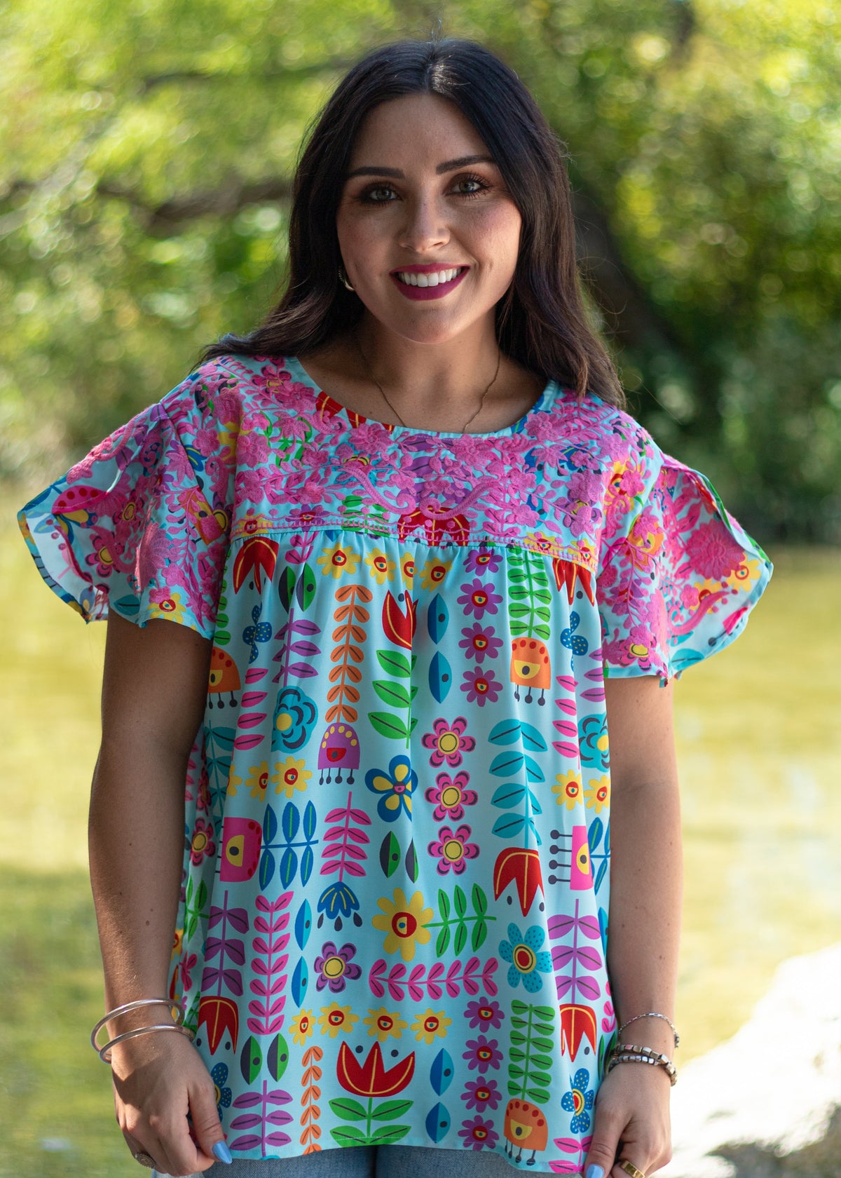 Woman with dark hair standing in a front profile position wearing the Butterfly Top in a lifestyle scene. The focal point of this image is the front detailed embroidery and floral graphics. The model paired this top with light-wash denim pants. 