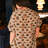 Woman with dark hair standing in a back profile position wearing the Calamity Top in a lifestyle scene. The focal point of this image is the cowboy hat graphics and visibility of cuff and collar. The model paired this top with the same graphic pants. 