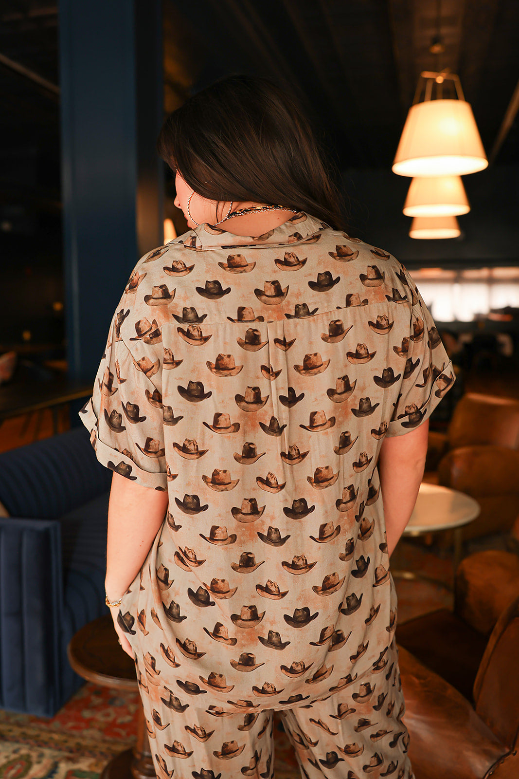 Woman with dark hair standing in a back profile position wearing the Calamity Top in a lifestyle scene. The focal point of this image is the cowboy hat graphics and visibility of cuff and collar. The model paired this top with the same graphic pants. 