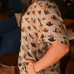 Woman with dark hair standing in a side profile position wearing the Calamity Top in a lifestyle scene. The focal point of this image is the cowboy hat graphics. The model paired this top with the same graphic pants. 