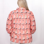 Woman with blonde hair standing in a back profile position wearing the Caroline Top against a white background. The focal point of this image is the boot graphics against pink shirt. The model paired this top with white pants. 
