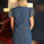 Woman with blonde hair standing in a back profile position wearing the Caylee Dress in a lifestyle scene. The focal point of this image is the denim fabric and ruffles sleeves with cream floral embroidery. The model paired this dress with brown boots. 