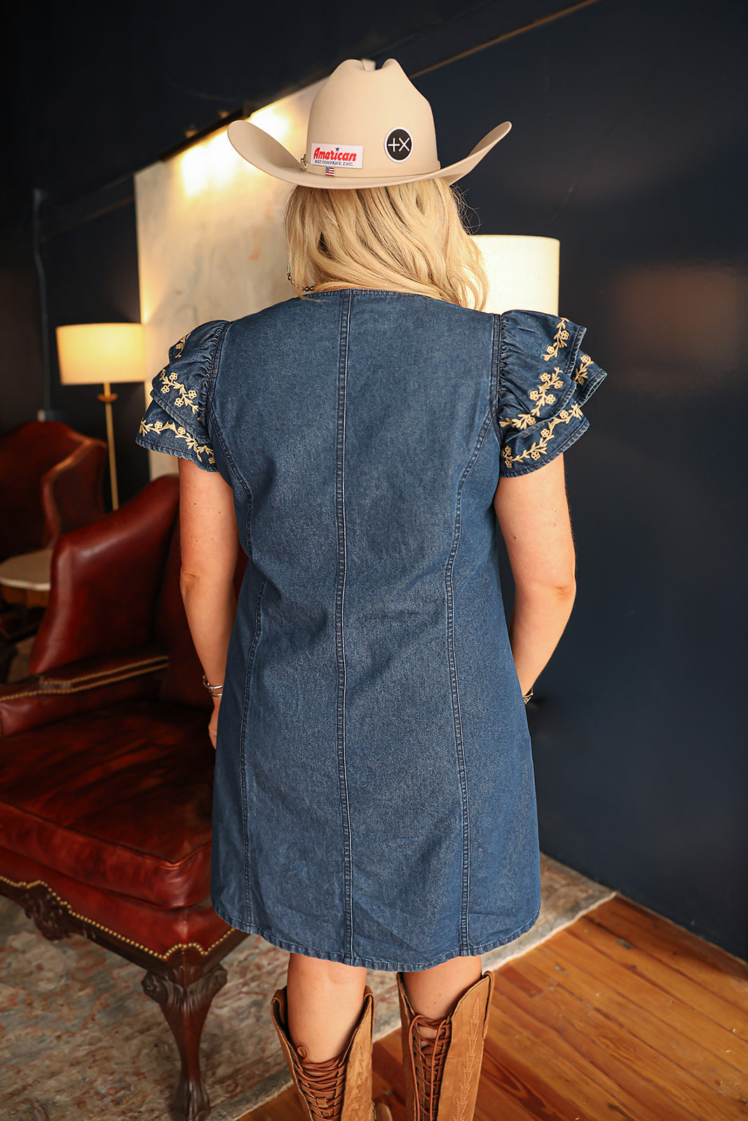 Woman with blonde hair standing in a back profile position wearing the Caylee Dress in a lifestyle scene. The focal point of this image is the denim fabric and ruffles sleeves with cream floral embroidery. The model paired this dress with brown boots. 