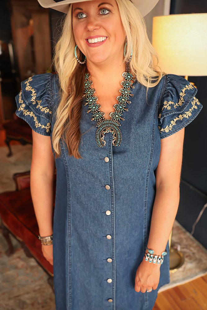 Woman with blonde hair standing in a front profile position wearing the Caylee Dress in a lifestyle scene. The focal point of this image is the buttons down front of dress, ruffled sleeves, and cream floral embroidery. 
