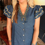 Woman with blonde hair standing in a front profile position wearing the Caylee Dress in a lifestyle scene. The focal point of this image is the buttons down front of dress, ruffled sleeves, and cream floral embroidery. 