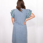 Woman with dark hair standing in a back profile position wearing the Caylee Dress against a white background. The focal point of this image is the denim dress and back of ruffle sleeves with green and yellow embroidery. 