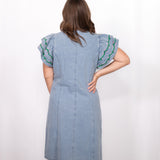 Woman with dark hair standing in a back profile position wearing the Caylee Dress against a white background. The focal point of this image is the denim dress and back of ruffle sleeves with green and yellow embroidery. 
