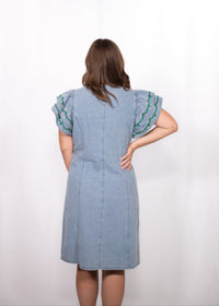 Woman with dark hair standing in a back profile position wearing the Caylee Dress against a white background. The focal point of this image is the denim dress and back of ruffle sleeves with green and yellow embroidery. 