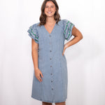 Woman with dark hair standing in a front profile position wearing the Caylee Dress against a white background. The focal point of this image is the dresses entirety (buttons, denim, ruffle sleeve). 