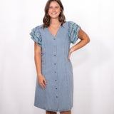 Woman with dark hair standing in a front profile position wearing the Caylee Dress against a white background. The focal point of this image is the dresses entirety (buttons, denim, ruffle sleeve). 