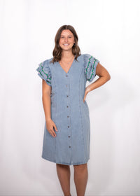 Woman with dark hair standing in a front profile position wearing the Caylee Dress against a white background. The focal point of this image is the dresses entirety (buttons, denim, ruffle sleeve). 