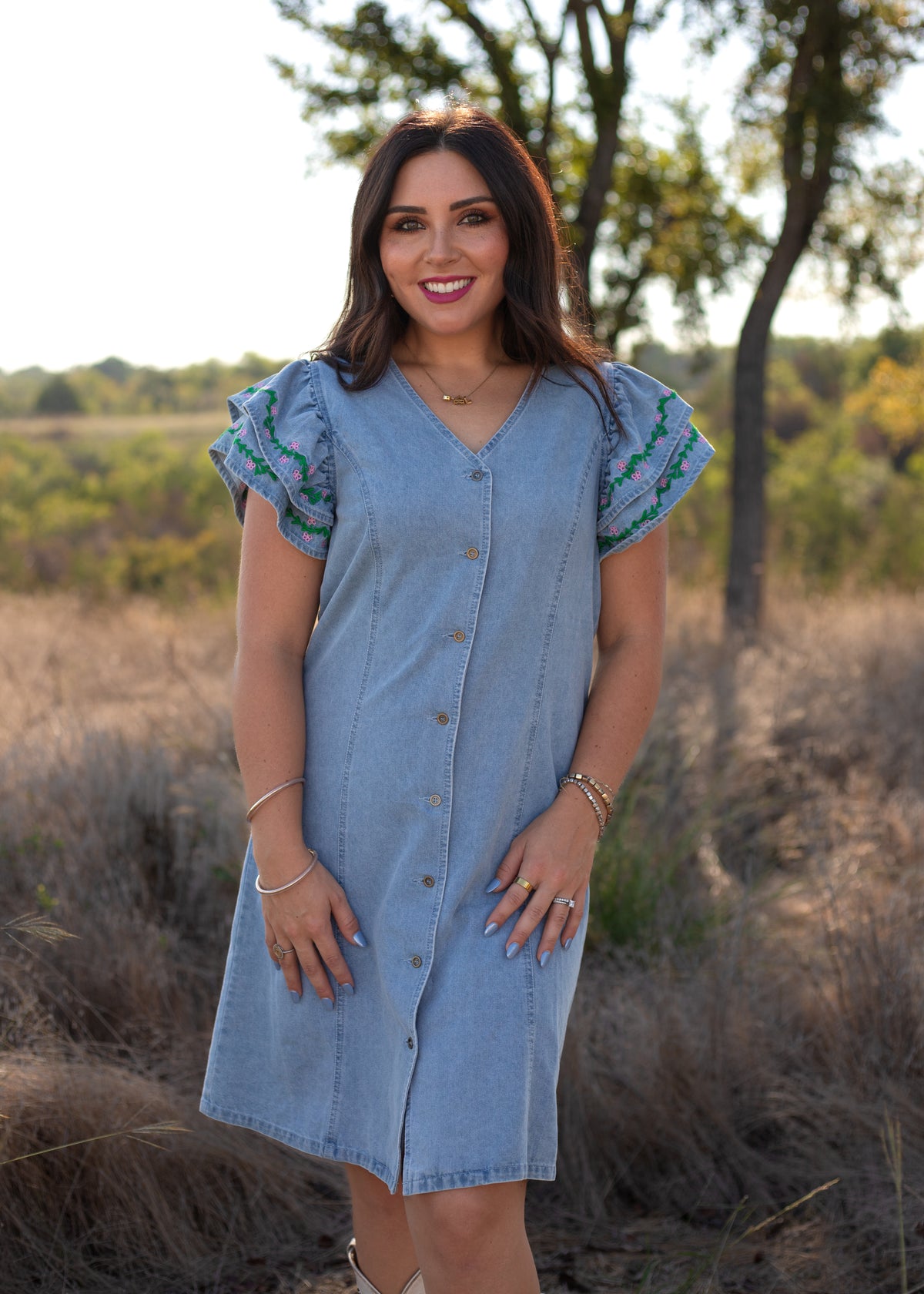 Woman with dark hair standing in a front profile position wearing the Caylee Dress in a lifestyle scene. The focal point of this image is the buttons down the front of dress as well as a v-neckline. 