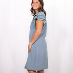 Woman with dark hair standing in a side profile position wearing the Caylee Dress against a white background. The focal point of this image is the side of denim dress and ruffle sleeves with green/yellow embroidery. 
