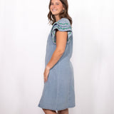 Woman with dark hair standing in a side profile position wearing the Caylee Dress against a white background. The focal point of this image is the side of denim dress and ruffle sleeves with green/yellow embroidery. 