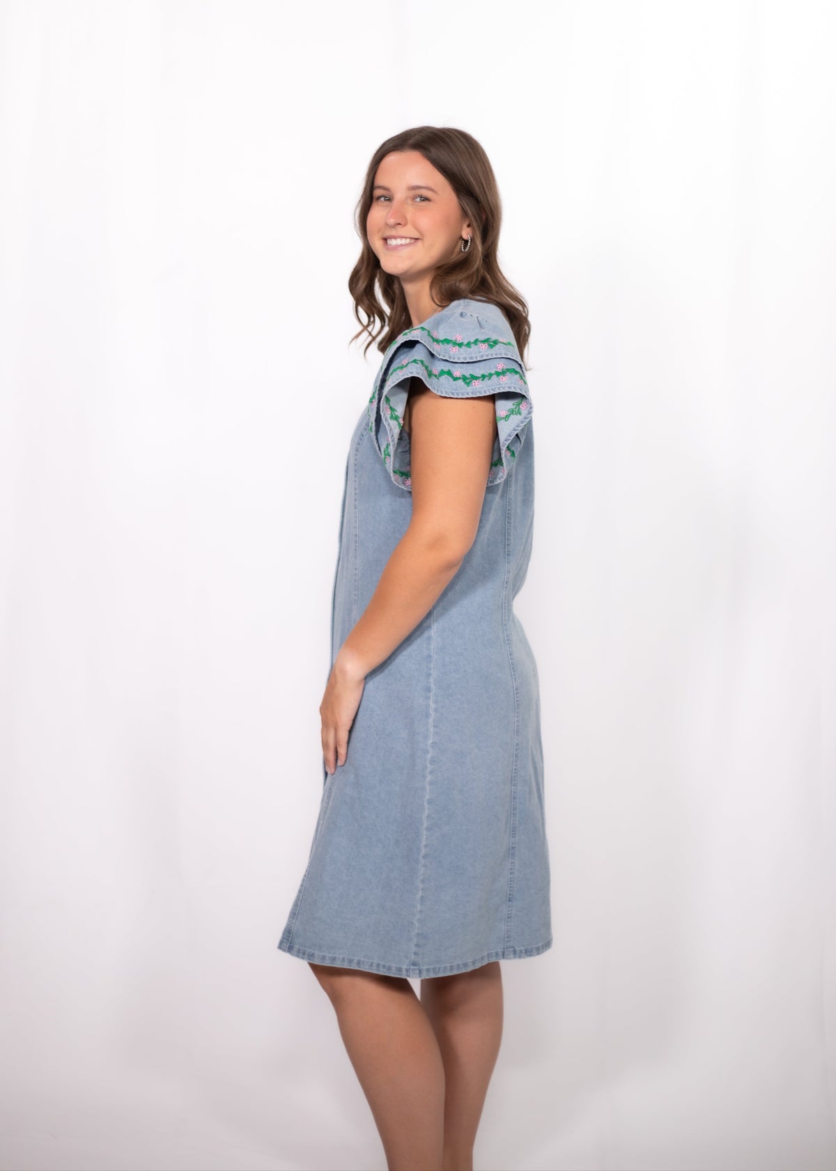 Woman with dark hair standing in a side profile position wearing the Caylee Dress against a white background. The focal point of this image is the side of denim dress and ruffle sleeves with green/yellow embroidery. 