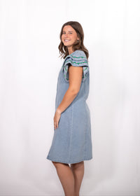 Woman with dark hair standing in a side profile position wearing the Caylee Dress against a white background. The focal point of this image is the side of denim dress and ruffle sleeves with green/yellow embroidery. 