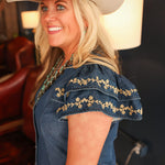 Woman with blonde hair standing in a side profile position wearing the Caylee Dress in a lifestyle scene. The focal point of this image is the ruffled sleeves with cream floral embroidery. 