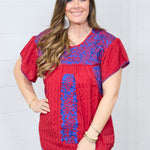 Woman with dark hair standing in a front profile position wearing the Chelsea Top against a white background. The focal point of this image is the blue floral embroidery across chest and down the top. The model paired this top with dark-wash denim shorts.