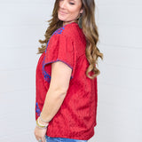Woman with dark hair standing in a side profile position wearing the Chelsea Top against a white background. The focal point of this image is the blue embroidery on shoulders, loose sleeves, buck-stitch on cuff. The model paired this top with dark-wash denim shorts.