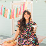 Woman with dark hair in a lifestyle scene wearing the Claire Dress. The background of image is a backyard scene with emphasis on pops of colors. 