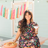 Woman with dark hair in a lifestyle scene wearing the Claire Dress. The background of image is a backyard scene with emphasis on pops of colors. 