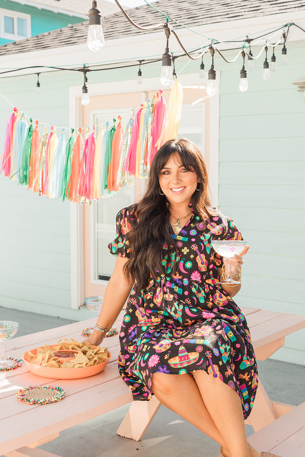 Woman with dark hair in a lifestyle scene wearing the Claire Dress. The background of image is a backyard scene with emphasis on pops of colors. 