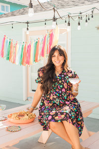 Woman with dark hair in a lifestyle scene wearing the Claire Dress. The background of image is a backyard scene with emphasis on pops of colors. 