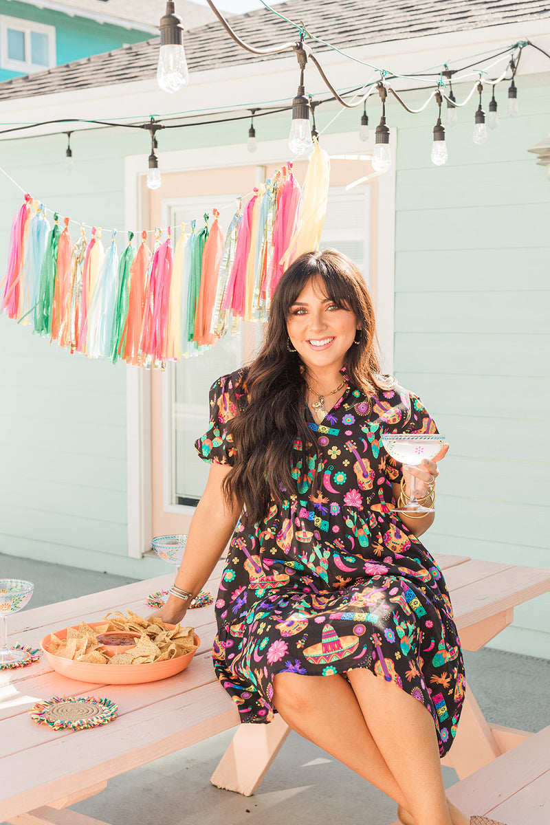 Woman with dark hair in a lifestyle scene wearing the Claire Dress. The background of image is a backyard scene with emphasis on pops of colors. 