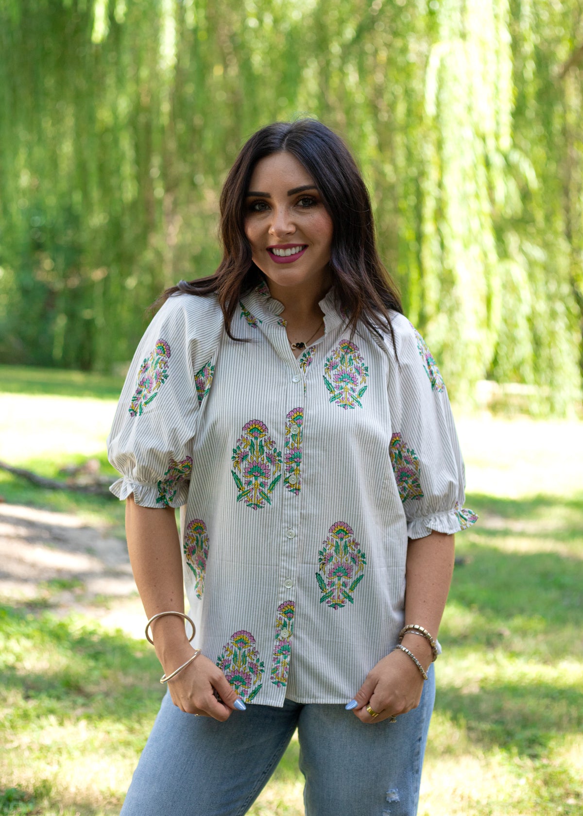 Woman with dark hair wearing the Courtney Top in a front facing position with lifestyle background. The background  is heavy in greenery, and the model has the top paired with light-wash denim jeans. 