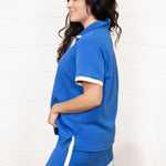 Woman with dark hair standing in a side profile position wearing the Daisy Top and Jane Bottoms set against a white background. The focal point of this image is seeing the design of the fabric from the side as well as judgement on length of skirt. 