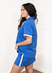 Woman with dark hair standing in a side profile position wearing the Daisy Top and Jane Bottoms set against a white background. The focal point of this image is seeing the design of the fabric from the side as well as judgement on length of skirt. 