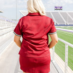 Woman with blonde hair standing in a back profile position wearing the Daisy Top and Jade Skirt at a football field. The focal point of this image is the intricate fabric detail on the back of top and skirt as well as the striping on cuff. 